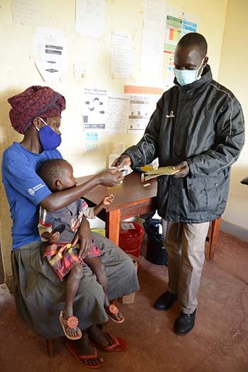 A health work hands a dose of ivermectin to a seated woman holding a child.