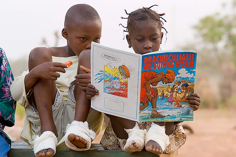 Photo of a young girl and young boy with their feet wrapped in gauze sitting on a bench reading a comic book called Dracunculiasis or Guinea worm.