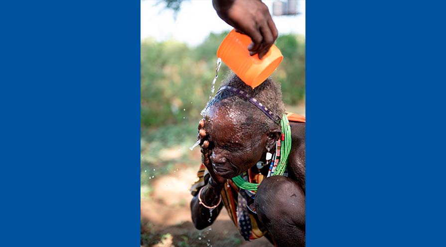 Photo of a woman kneeling as she receives help rinsing her face with water outdoors in her village.