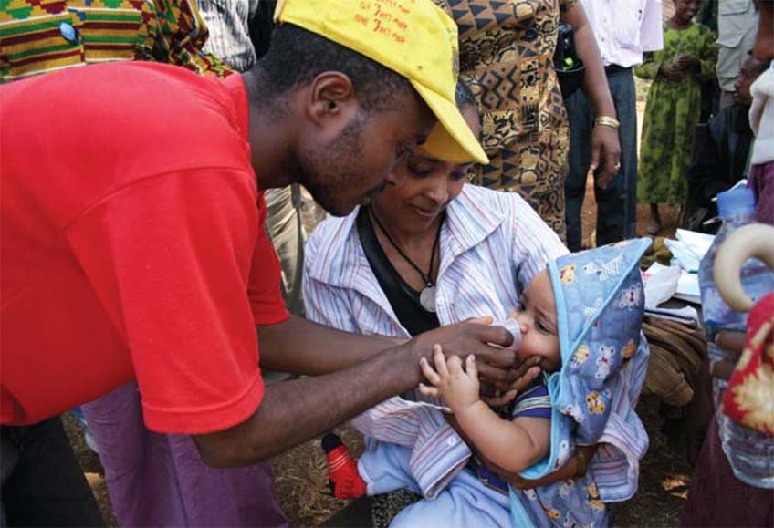 A male worker wearing a red shirt and yellow caps gives medication to a baby being held by mom.